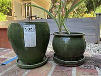 Front view of two green glazed ceramic plant pots with attached saucers, the larger one on the left and smaller one on the right with a plant in each pot
