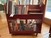 Red low two-tier children's library cart fully loaded with books on wooden floor