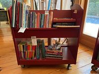 Front view of red Land of Nod children's library cart filled with various children's books on upper and lower shelves.