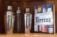 Three polished metal cocktail mixers and one cocktail recipe book shown from the front with the book cover visible.