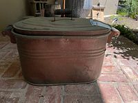 Front view of the copper tub with lid on a brick floor outdoors, showing side handles and tarnishing on the copper surface.