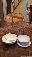 Two-tier metal pastry rack standing behind two ceramic bowls and a cheese cutter placed on a stone floor.