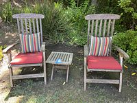 Pair of weathered teak wood outdoor chairs with two red seat cushions and two multicolored striped back cushions flanking a small teak side table with blue tape.