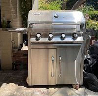 Front view of the stainless steel Turbo STS gas grill showing four control knobs, double-door cabinet, and propane tank on side shelf.