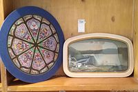 Two wall art pieces on a wooden shelf: round blue-framed painting of rose window and curved rectangular framed waterfall print.