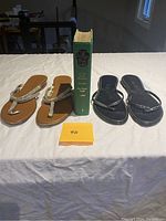 Two pairs of Toscanella sandals and a green covered book on a white cloth surface.