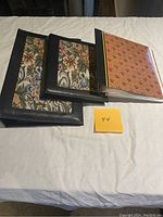 Three photo album books arranged side by side showing front covers; two with floral needlework and one with patterned orange cover.