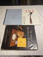 Photo showing three books: 'The Book of Canadian Antiques' with an antique telephone on the cover, a spiral-bound 'Some Brass Hardware' book with a gray cover, and 'ART' Canadian Art book with a sculpture on the cover.