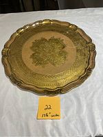 Front view of round gold toned platter with embossed floral ornamental design and scalloped edge, placed on white cloth with a note indicating 17.25 inches wide.