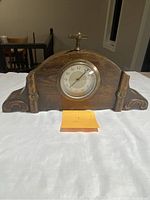Front view of the antique wooden cabinet clock showing round clock face with Arabic numerals and floral carved wood decoration on curved clock body.