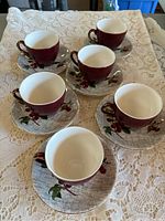 Set of six burgundy teacups with matching saucers displayed on lace tablecloth.