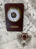 Birks barometer mounted on wood with blank nameplate and Villeroy & Boch heart-shaped paperweight clock beside it on lace fabric.