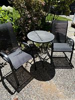 Outdoor bistro set with round table and two chairs, placed outside on a patio with plants nearby.