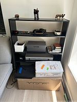 Black bookshelf containing various items on shelves, including small animal figurines, toy cars, boxes, books, and a briefcase. Adjacent to a gray couch and near a window.