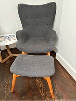 Front view of gray upholstered chair with matching ottoman on dark wood floor near white wall and corner table.