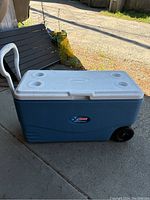 Closed blue Coleman cooler with white lid, cup holders on the lid and wheels on one side.