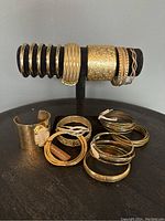 Photo of gold tone costume bangles and bracelets displayed on a black jewelry stand and surface. Various widths and styles shown including wide cuff and multiple slim bangles, some textured or etched.