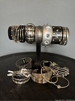 Photo showing an assortment of silver tone costume jewelry bangles including various styles and designs on black stand and scattered on surface.