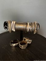 The photo shows a black jewelry display stand holding a collection of silver tone costume jewelry including thin and thick bangles with various textures, and multiple bracelets. In front of the display are two cuff bracelets and one square-link bracelet.
