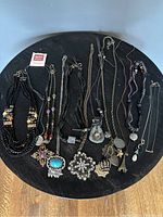 Photo showing an assortment of 11 costume jewelry necklaces arranged on a black round surface. Necklaces feature various designs including beaded, braided fabric, chains, and different pendants such as turquoise stone, floral rhinestones, crosses, and coins.