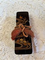 Closed view of rectangular black lacquer fumbako box decorated with gold flowing water and floral designs, tied with a reddish corded tassel.