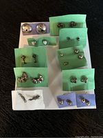 Photo showing 11 pairs of small stud earrings on different colored paper bases, silver-tone metal, varied shapes including hearts, bows, spheres, and geometric.
