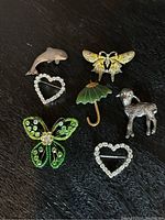 Photo showing seven brooches arranged on a dark surface: silver lamb, jade umbrella, dolphin, two rhinestone hearts, green rhinestone butterfly, and bee/moth with yellow wings.