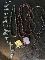 Photo of multiple necklaces and earrings laid out on dark surface, showing variety of bead types including amethyst, hematite, pearl, clear crystal, and garnet/dark red stones.