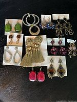 Photo shows twelve pairs of assorted earrings in multiple styles and colors placed on a black surface.