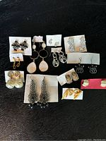 Photo displaying twelve pairs of assorted earrings arranged on a black surface on various paper cards, showing a variety of styles including studs, hoops, and dangles in silver, gold, and pearl-like finishes.