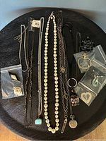 Top-down image showing assorted necklaces and pendants laid out on a dark circular table surface, including a pearl necklace, multiple chain necklaces, and several pendants some in small plastic bags.