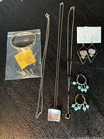 Six assorted pieces of silver-marked jewelry displayed against a black background: a bangle bracelet in plastic bag, two necklaces with rectangular pendants, two pairs of earrings with floral and bead designs.