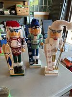 Photo showing three wooden nutcracker figurines dressed as skiers or snowboarders, placed on a table with a garage background.