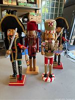 Four wooden nutcrackers on a table, two pirates with black hats and gold trim, one candy maker with peppermint theme, and one voyager with red hat and checkered coat.