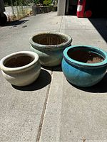 Photo showing three outdoor ceramic planters in white, light green, and turquoise blue placed on concrete.