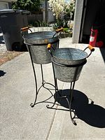 Pair of two galvanized metal planters on black metal stands with wooden handles placed outdoors on concrete.