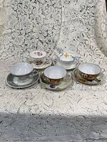 Five assorted porcelain teacup and saucer sets placed on white lace fabric, displaying various floral patterns and gold trim on rims and handles.
