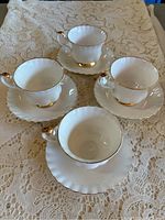 Four white porcelain Royal Albert teacups with gold trim around rims and handles along with matching saucers displayed on lace tablecloth.