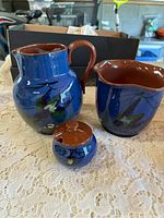 Three-piece Torquay pottery set: jug, pitcher with scalloped rim, and small covered jar with floral decoration on blue glaze.