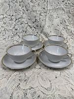 Four white porcelain teacups and saucers arranged on lace fabric, showing gold trim detail around edges.