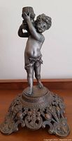 Full front view of a standing cast iron cherub figure holding a lamp holder on an ornate base.