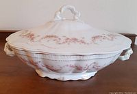 Side profile of the covered oval dish showing pink floral garlands and gold trim on lid, with white porcelain handles and scalloped edges.