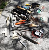 Wide photo showing assortment of used hand tools spread on pavement including hammers, crow bars, cutting discs, scrapers, and other hand tools
