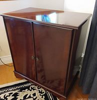 Front and side angle view of closed Bombay Company cherry wood TV cabinet with two doors and brass-tone handles.