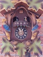 Front close-up of the cuckoo clock showing carved birds, detailed leaves, clock face with roman numerals and dancing couple figurines