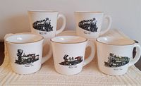 Five white ceramic coffee mugs arranged in two rows featuring detailed locomotive train illustrations in black, red, and gold. Gold trim visible on rims. Mugs have cylindrical shape with wide handles.