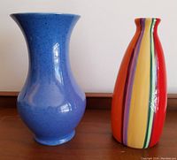Two vases side by side on a wooden surface, showing the powder blue antique William Moorcroft vase and the multi-colour retro striped vase