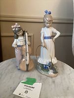 Front view of two NAO by Lladro figurines - girl playing cello and girl with dog and hoop, positioned on a marble table.
