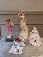 Four ceramic figurines arranged on marble surface including Royal Worcester 'Wednesday's Child' in pink dress, Royal Doulton lady with clover dress, Staffordshire swan, and seated girl in white dress.