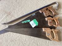 Three hand saws with wooden handles and varying blade lengths stacked on floor along with one metal crowbar showing wear.